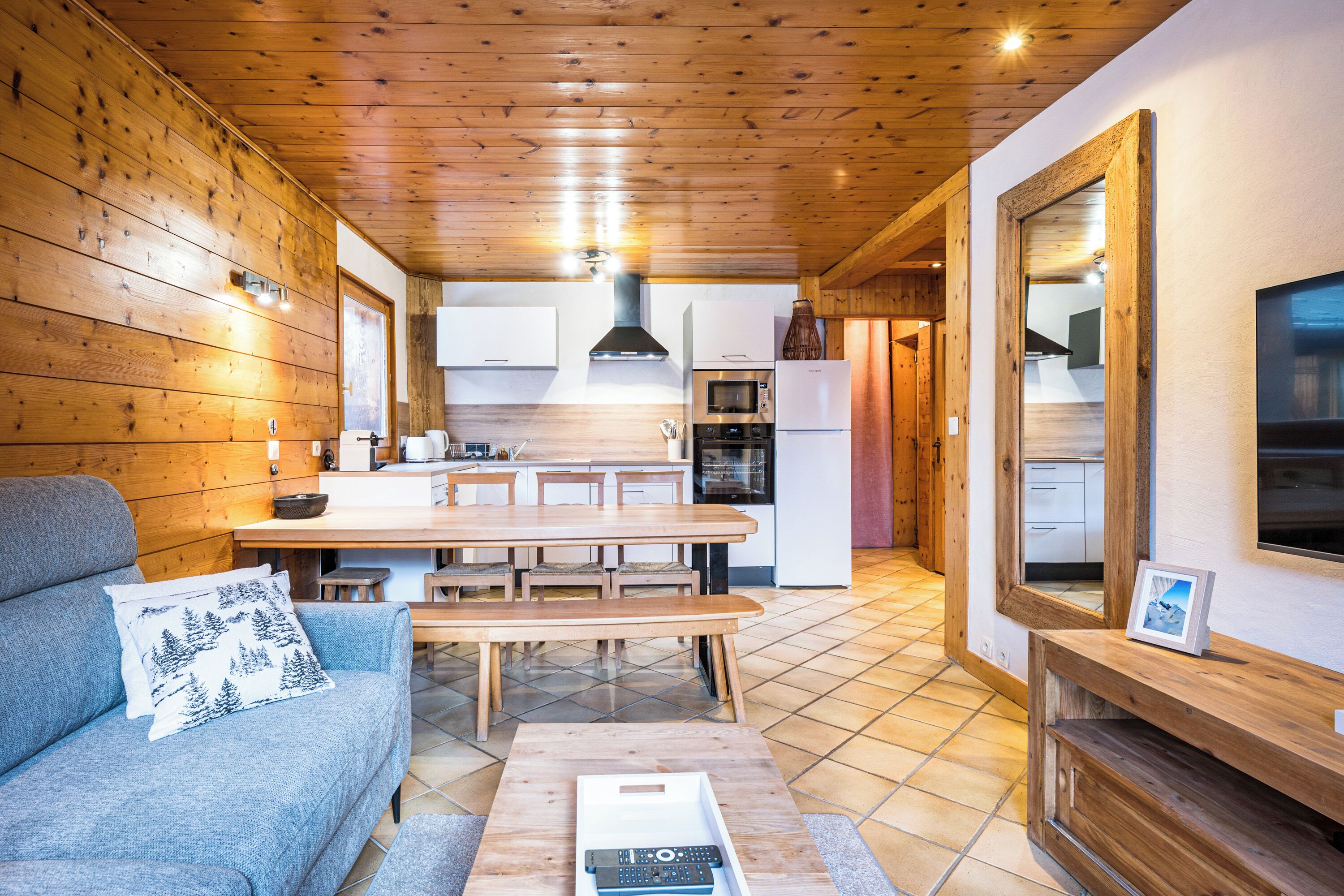 A cozy cabin interior with a wooden ceiling, a dining area with a table and benches, a kitchen with modern appliances, and a large mirror on the wall.