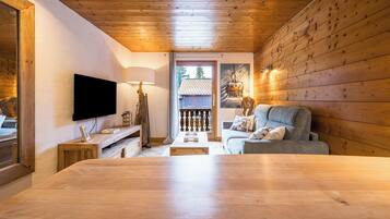 A cozy room with a wooden ceiling, a flat-screen TV mounted on the wall, a grey sofa with cushions, and a wooden table in the center.