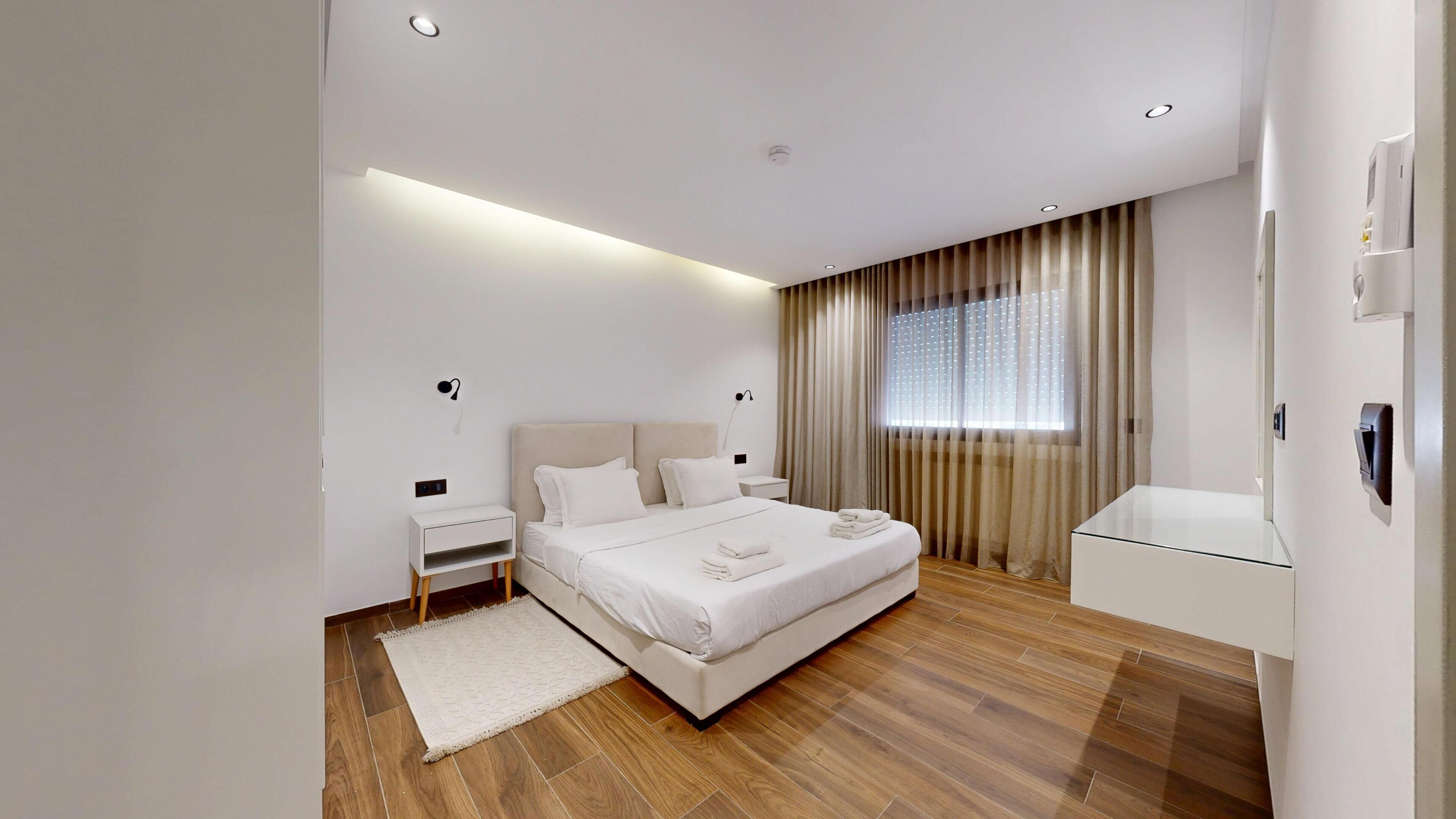 Una habitación de hotel moderna con una cama grande, mesitas de noche, tocador y ventana con cortinas.
