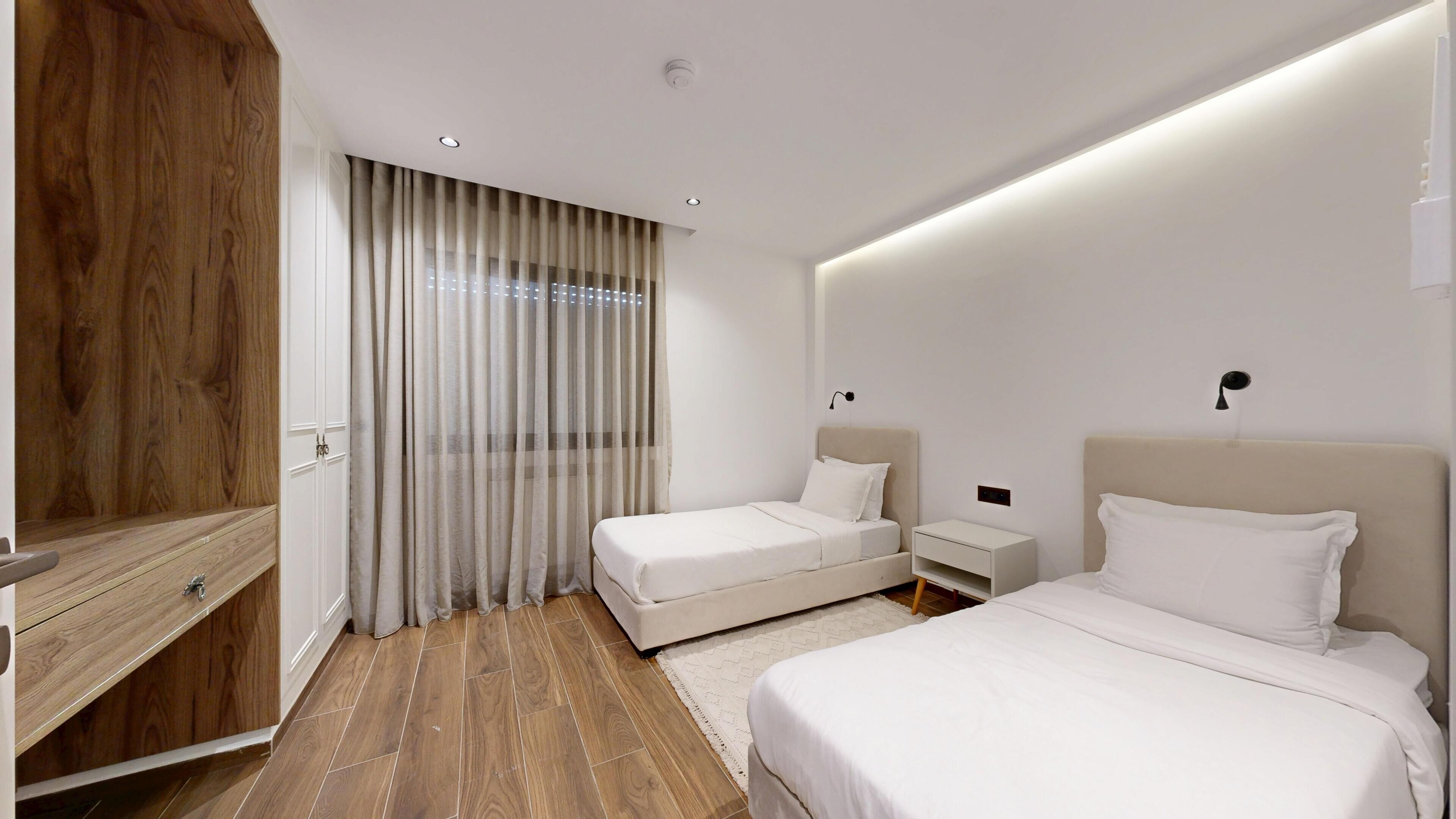 A modern hotel room with two beds, a wooden wardrobe, a large window with curtains, and a nightstand.