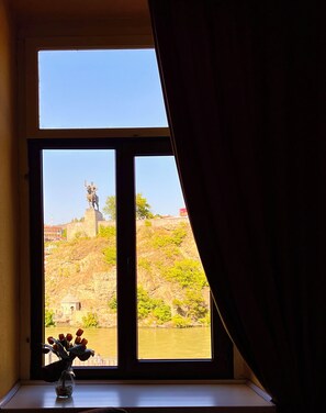 A window with a view of a statue on a hill, a vase with flowers on a windowsill, and a curtain partially drawn. - Old Ubani apartment with two bedrooms and a living room,Private WC and Bathroom. (T'bilisi)
