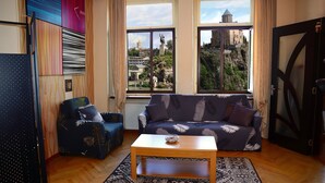 Een woonkamer met een bank, fauteuil en salontafel, grote ramen met uitzicht op een stadsbeeld en een standbeeld, en een kamerkleed op de vloer.