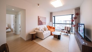 A modern living room with a sofa, coffee table, and TV, a large window with a city view, and a door leading to another room. - Tyms 3BR 2BA Apt  Gubei Fully Furnished (Shanghai)