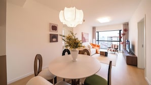 A modern living room with a round dining table, a potted plant, and a large window offering a city view. - Tyms 3BR 2BA Apt  Gubei Fully Furnished (Shanghai)