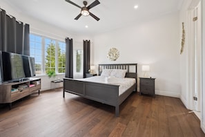 A bedroom with a bed, a TV, a ceiling fan, a nightstand, a lamp, and a window with curtains.
