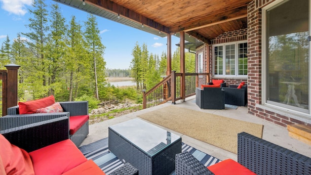 Un patio exterior techado con mobiliario de mimbre, una mesa de vidrio y vistas a un lago y un bosque.