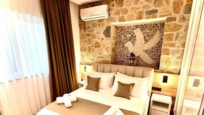 Habitación de hotel con pared de piedra, cama con ropa de cama blanca y marrón, un mosaico decorativo en la pared y una ventana con cortinas.