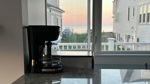A kitchen with a coffee maker on a marble countertop, a window with a view of a house and a dock. - Private Rye Beach getaway with 3 bedrooms across two units, ideal for couples or families. Enjoy private beach access, ocean views, fast Wi-Fi, and walk to Petey’s Famous Seafood—steps from the sand and coastal dining. (Rye)