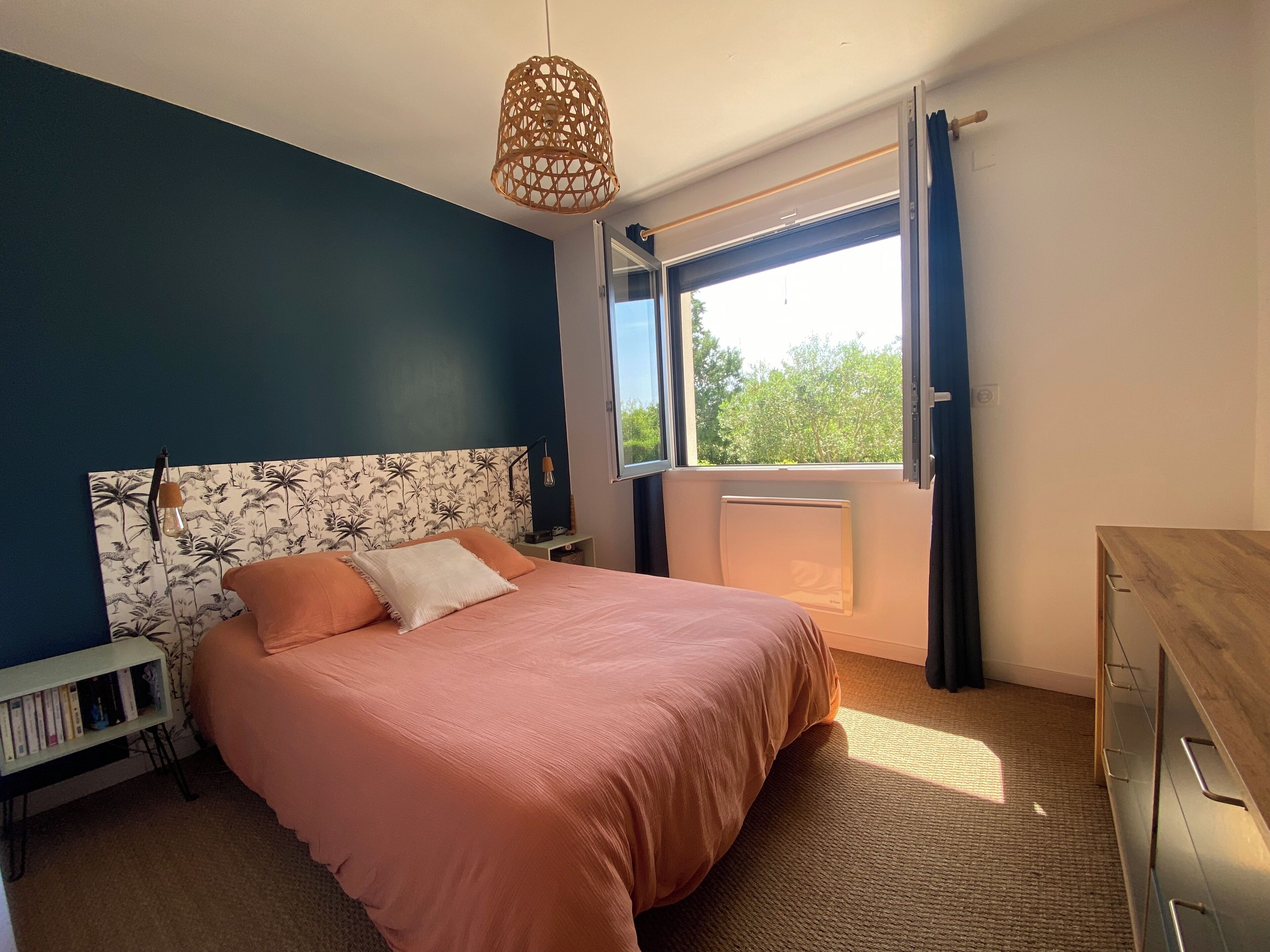 A bedroom with a bed, a nightstand, a dresser, a window with curtains, and a hanging light fixture.