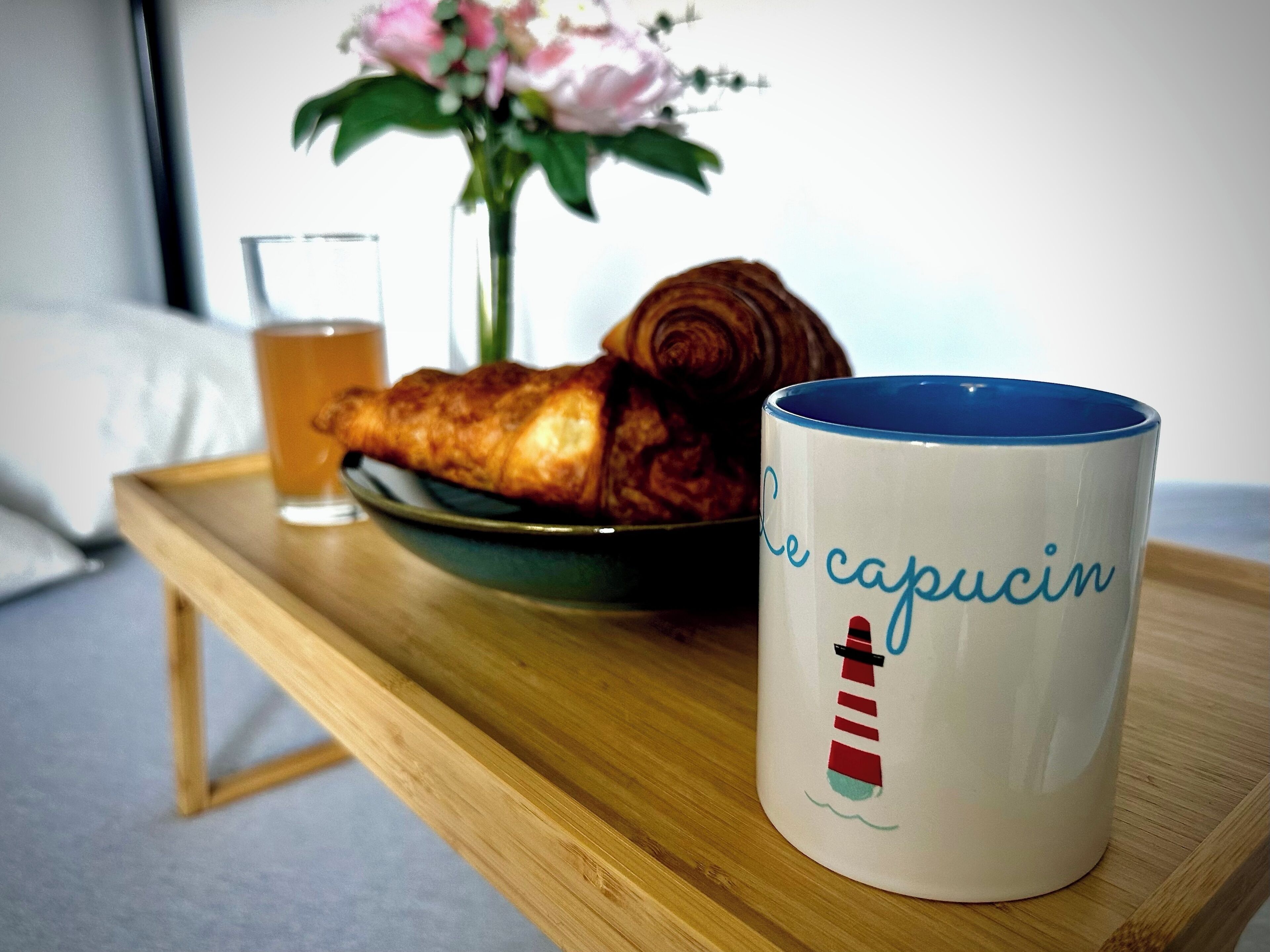 A wooden table with a croissant, a glass of tea, and a mug with a lighthouse design.