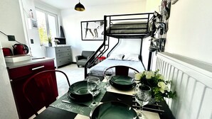 Quarto de hotel compacto com beliche, área de refeições com mesa posta para dois, pequena cozinha e janela com vista para área verde.