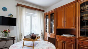 A dining area with a round table covered by a floral tablecloth, wooden cabinets with glass doors, a television mounted on the wall, and a window with curtains. - Lovely apartment for 6 people with WIFI and balcony (Riva Ligure)