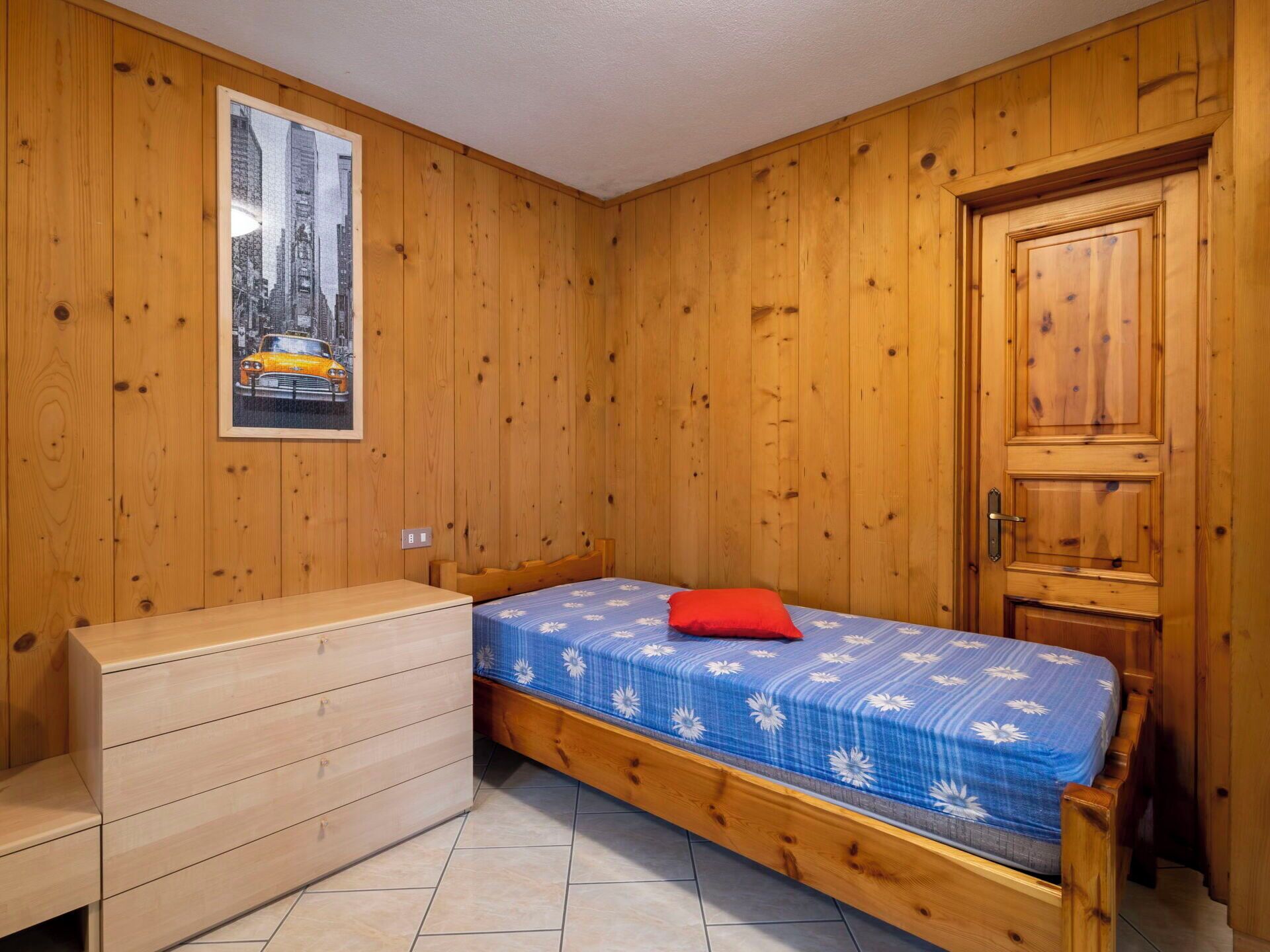 Una habitación pequeña con paredes de madera, una cama con colcha floral, un almohá de color rojo, un banco de madera y una puerta.