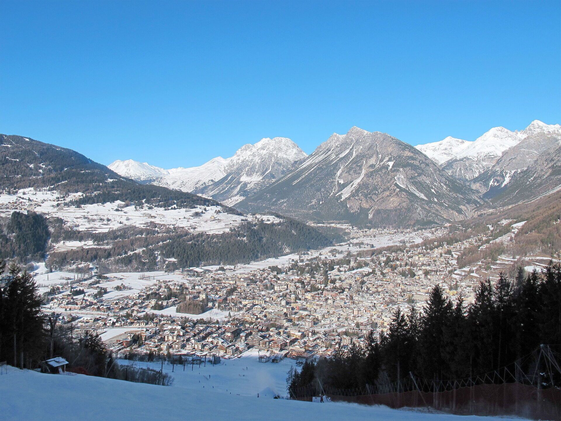 Une ville de montagne enneigée, avec un ensemble dense de bâtiments, une piste de ski et un ciel bleu dégagé.
