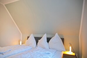 Ein ordentlich bezogenes Bett mit weißer Bettwäsche und Kissen, zwei Nachttische mit Lampen und eine schräge Decke.