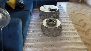 Una sala de estar moderna con un sofá seccional azul, dos mesitas redondas, un alfombra blanco de pelo alto y un amplio ventanal con vistas a muebles de exterior.