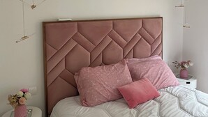 Un dormitorio con una cama grande, una mesita de noche blanca, un cojín decorativo rosa y un florero con flores.