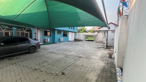 A paved courtyard with a car parked under a green canopy, buildings with blue walls, and flags on a wall. - Paskandy Hotel (Amasaman)