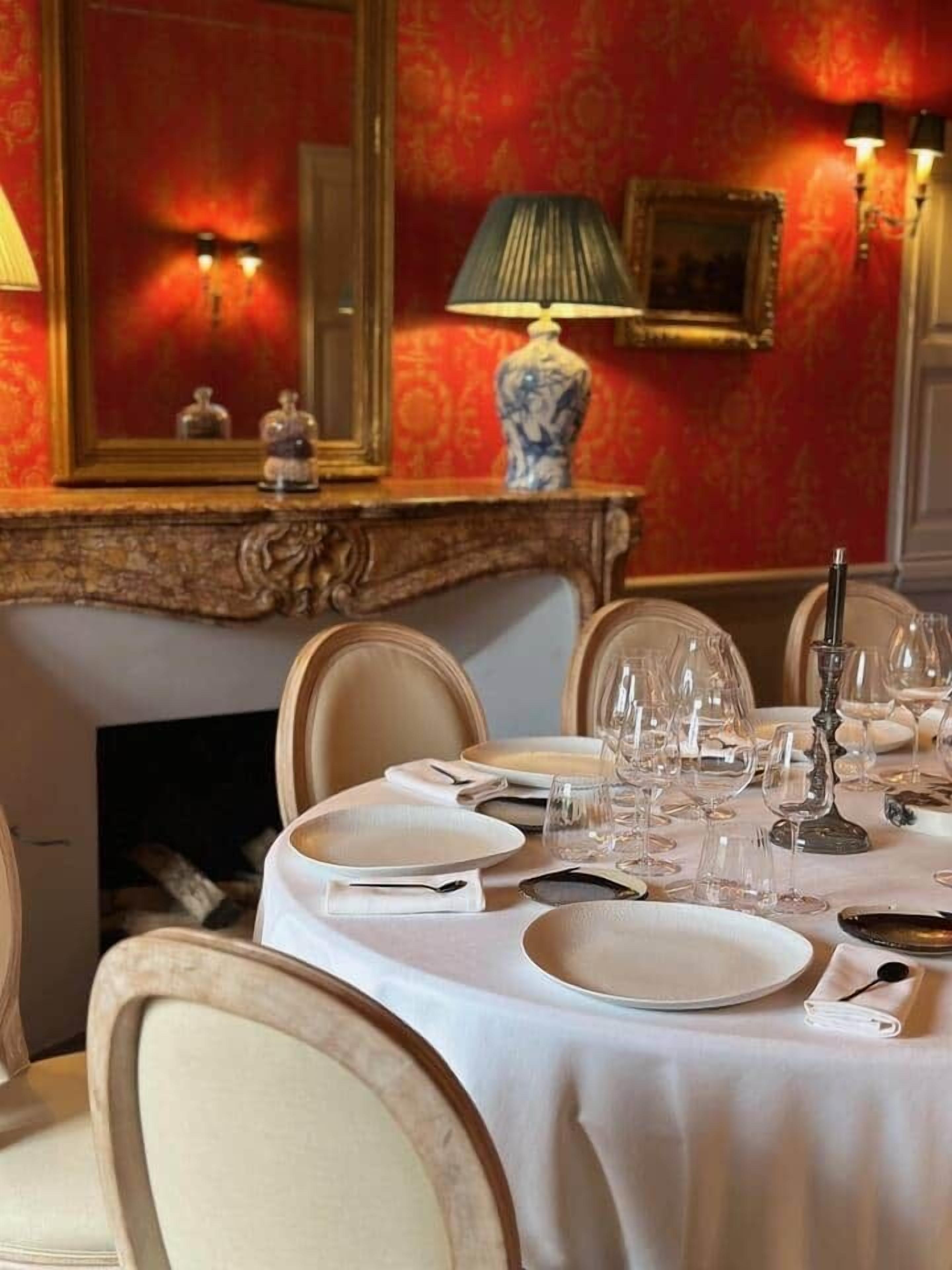 Una mesa de comedor dispuesta con platos, vasos y un candelabro, rodeada de sillas en una habitación con papel tapiz decorativo y un espejo.