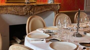 Una mesa de comedor dispuesta con platos, vasos y un candelabro, rodeada de sillas en una habitación con papel tapiz decorativo y un espejo.