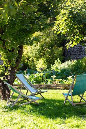 Deux fauteuils de jardin sont placés dans un espace verdoyant, avec un arbre et un mur de pierre en arrière-plan.