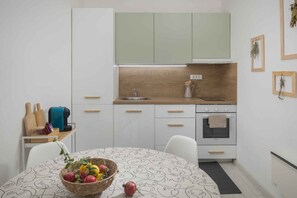 Eine kleine Küche mit weißen Schränken, einer Holz Arbeitsplatte, einem Spülbecken und einem Herd. Es steht ein Esstisch mit einer Korb voller Obst und einer Kaffeemaschine bereit.