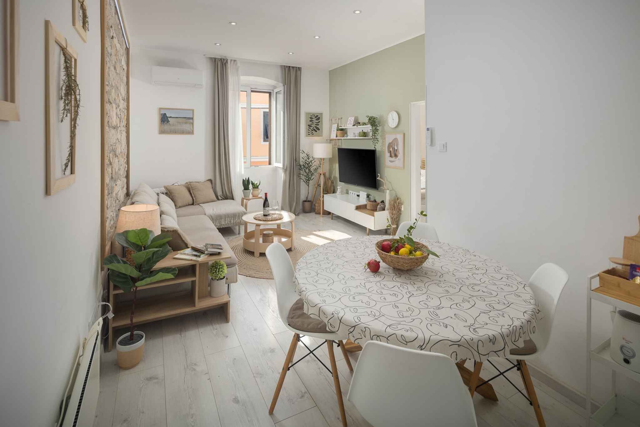 Ein modernes Wohnzimmer mit einer Couch, Couchtisch und Esstisch. Ein Fernseher steht auf einem Schrank und ein Fenster ist mit Vorhängen versehen.