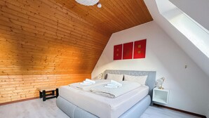 Quarto com teto inclinado, painéis de madeira, cama com roupa de cama branca, mesa de cabeceira e uma pequena janela.