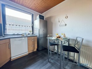 A compact kitchen with a dishwasher, refrigerator, and a dining table with chairs. - Apartment Uttied am Drangstweg (Cuxhaven)