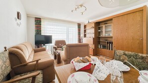 Uma sala de estar acolhedora com sofá, poltrona e mesa de jantar posta com pratos, xícaras e uma cesta de pão.