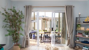 Una habitación con puerta corrediza de vidrio que da a un balcón, una planta en maceta, una estantería y una mesa con sillas.