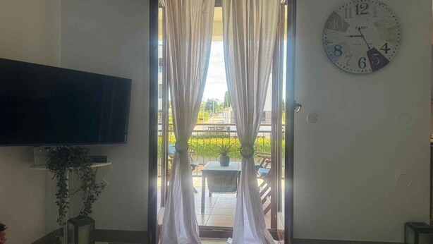 A room with a large flat-screen TV on the left, a wall clock, and a window with sheer curtains. - Holiday apartment with air conditioning and balcony (Poreč)