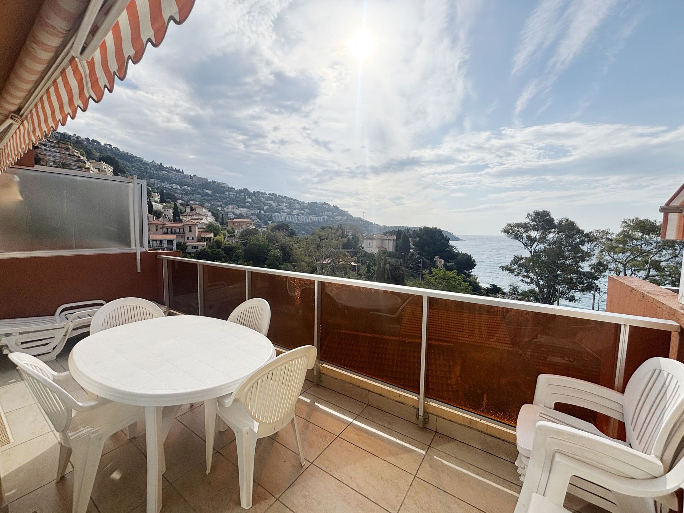 A balcony with a round table and chairs, overlooking a coastal town with buildings and trees.