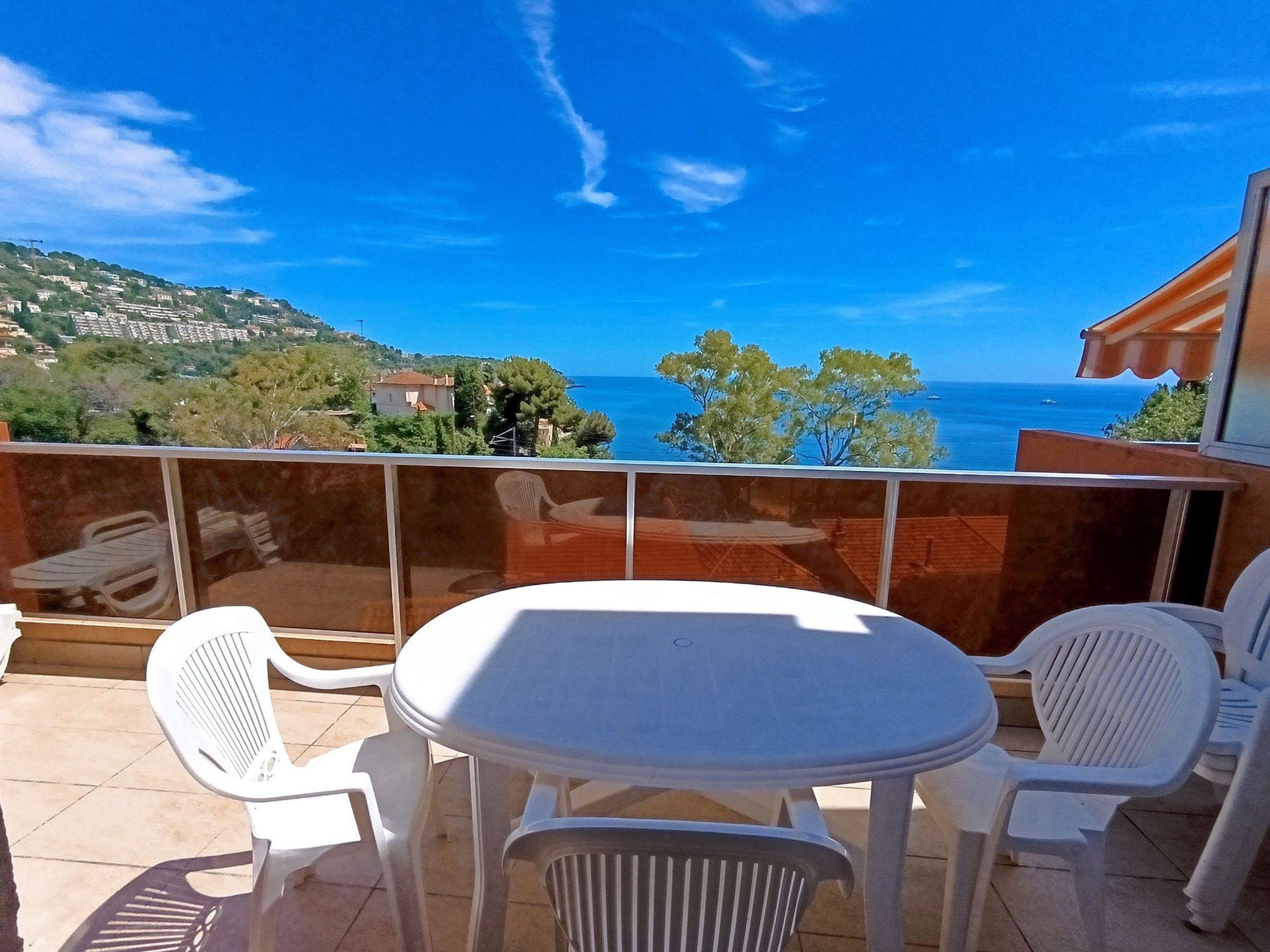 A balcony with a table and chairs, overlooking a coastal view with houses and trees.