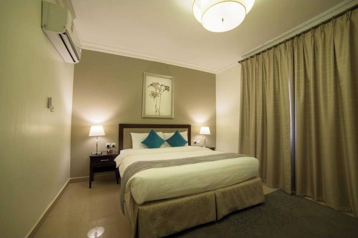 A hotel room with a bed, two bedside lamps, a nightstand, an air conditioning unit, a framed picture on the wall, and a window with curtains.