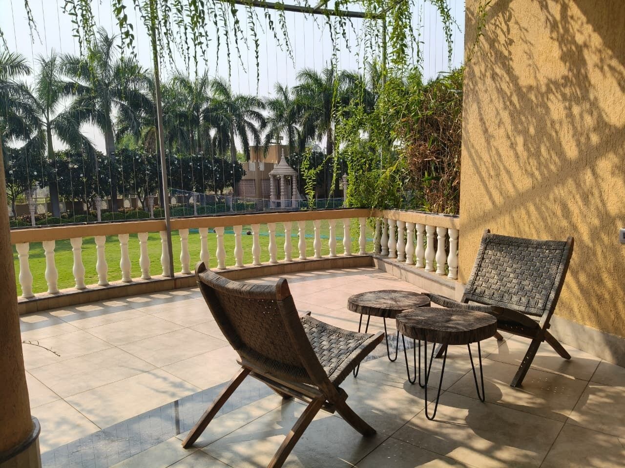 A balcony with two wicker chairs and a small round table, overlooking a garden with palm trees and a white fence.