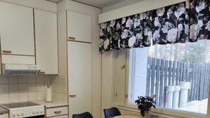 Una cocina compacta con armarios blancos, una estufa, una mesa de comedor con sillas y una ventana con cortina floral.