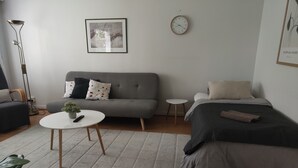 Una sala moderna con un sofá gris, una mesita redonda con una planta, una cama con cubierta oscura, una lámpara de pie y un cuadro enmarcado en la pared.