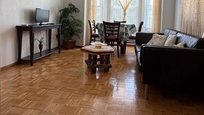 Un salon doté d’un parquet en bois disposé en motif «point de Hongrie», d’un canapé noir, d’une table à manger avec des chaises, d’un téléviseur posé sur un meuble et d’un luminaire fixé au plafond.
