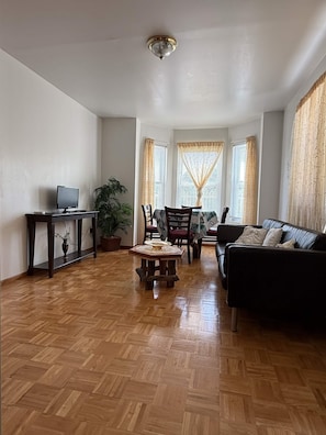 A living room with a wooden herringbone floor, a black sofa, a dining table with chairs, a TV on a stand, and a ceiling light fixture.