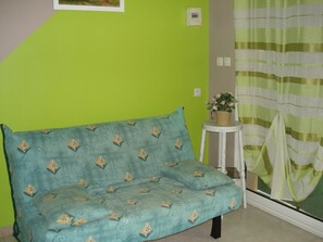 A teal sofa with a floral pattern, a small white stool with a potted plant, and a striped curtain. - SCI residence confort et loisirs (Gras)