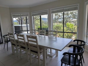 A dining area with a white table and chairs, large windows with blinds, and a view of greenery outside.