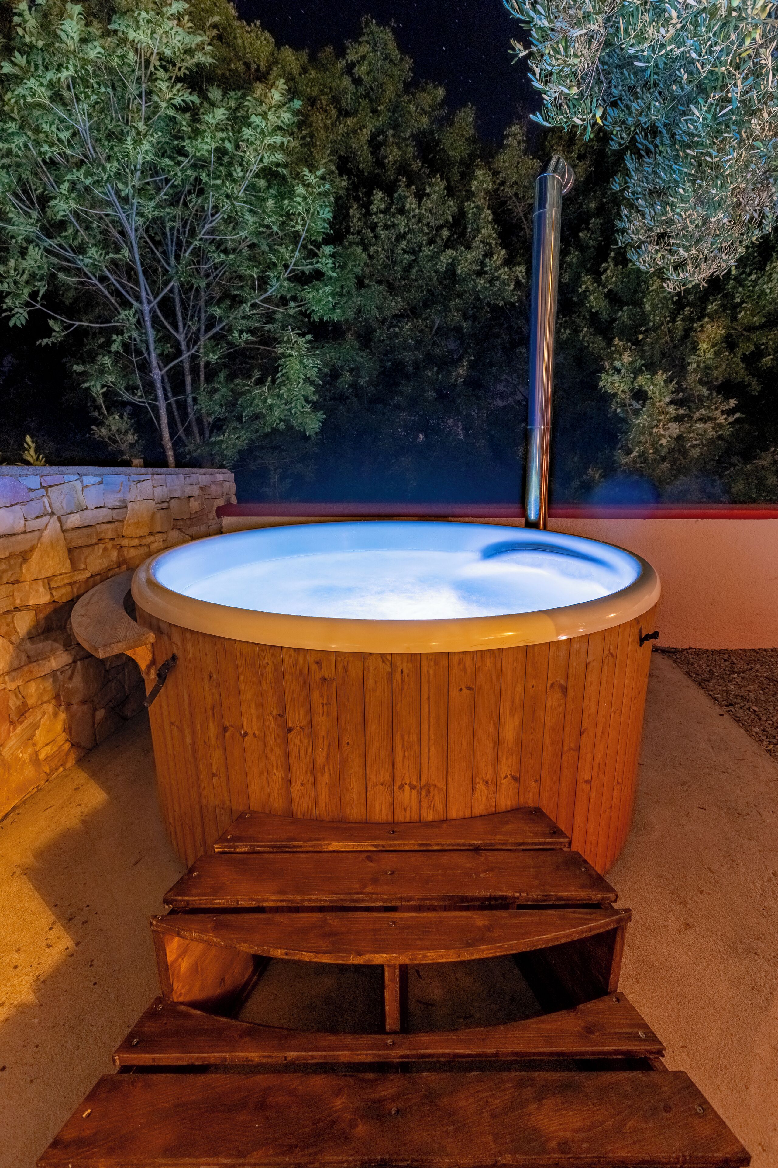 A wooden hot tub with a stone wall and steps leading up to it, surrounded by trees.