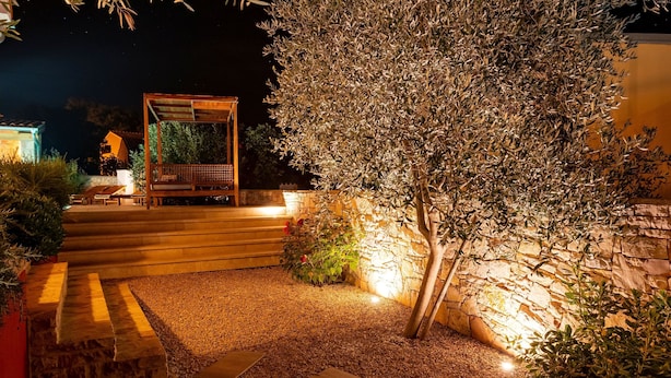 Zona exterior nocturna amb una pèrgola, escales que condueixen a una zona de seients i un camí de grava.