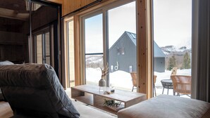 Een modern interieur met een bank, een houten bijzettafeltje en uitzicht op een berghut door grote ramen.