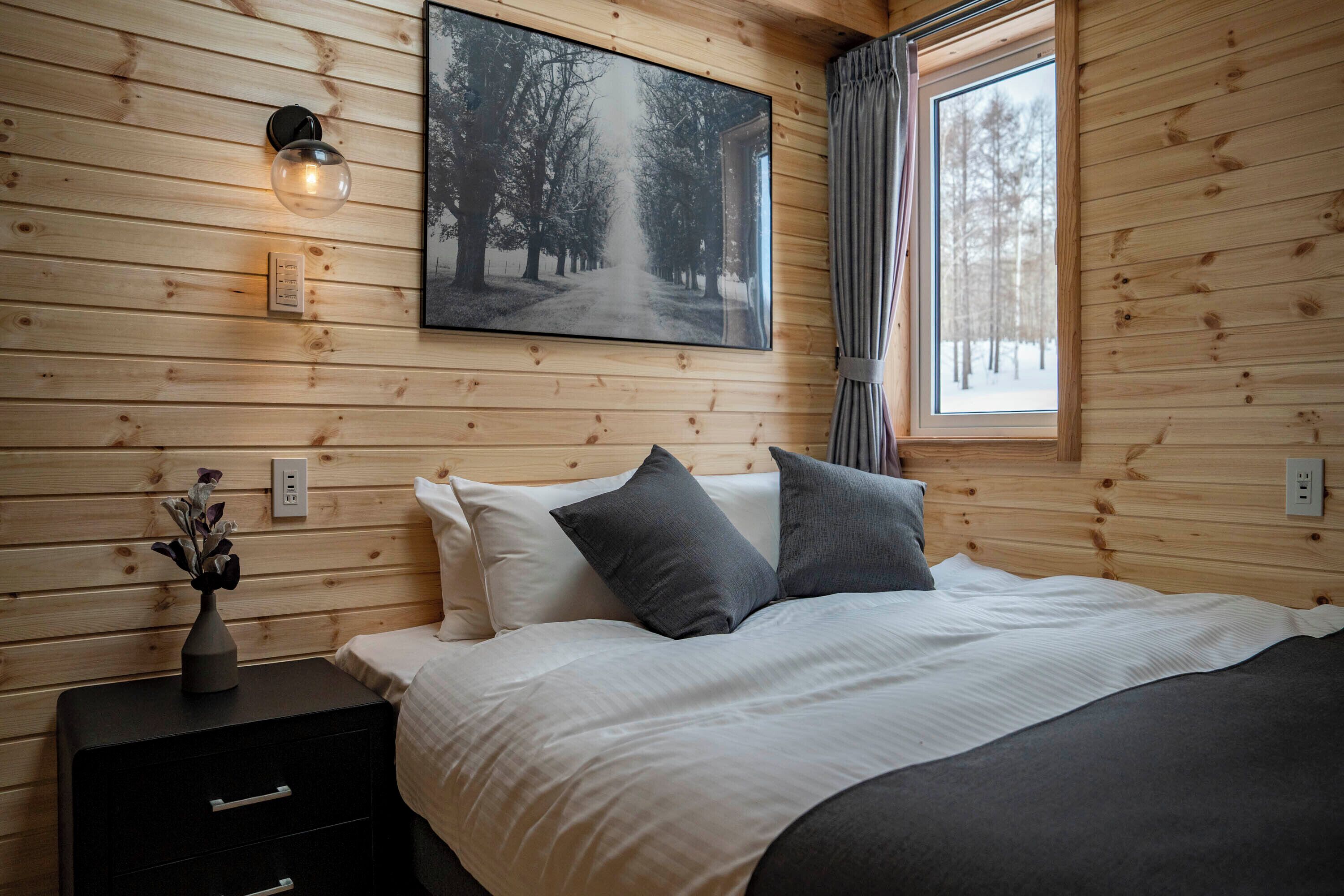 A wooden-paneled room with a bed, a nightstand, a wall-mounted light, and a window with curtains.
