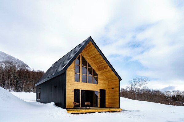 A wooden cabin with a large window, a porch, and a snowy landscape.