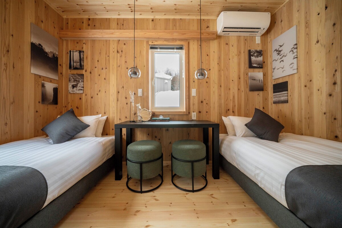 A cabin-style room with two single beds, a small table, and two stools. There is a window with a view of a snowy landscape.
