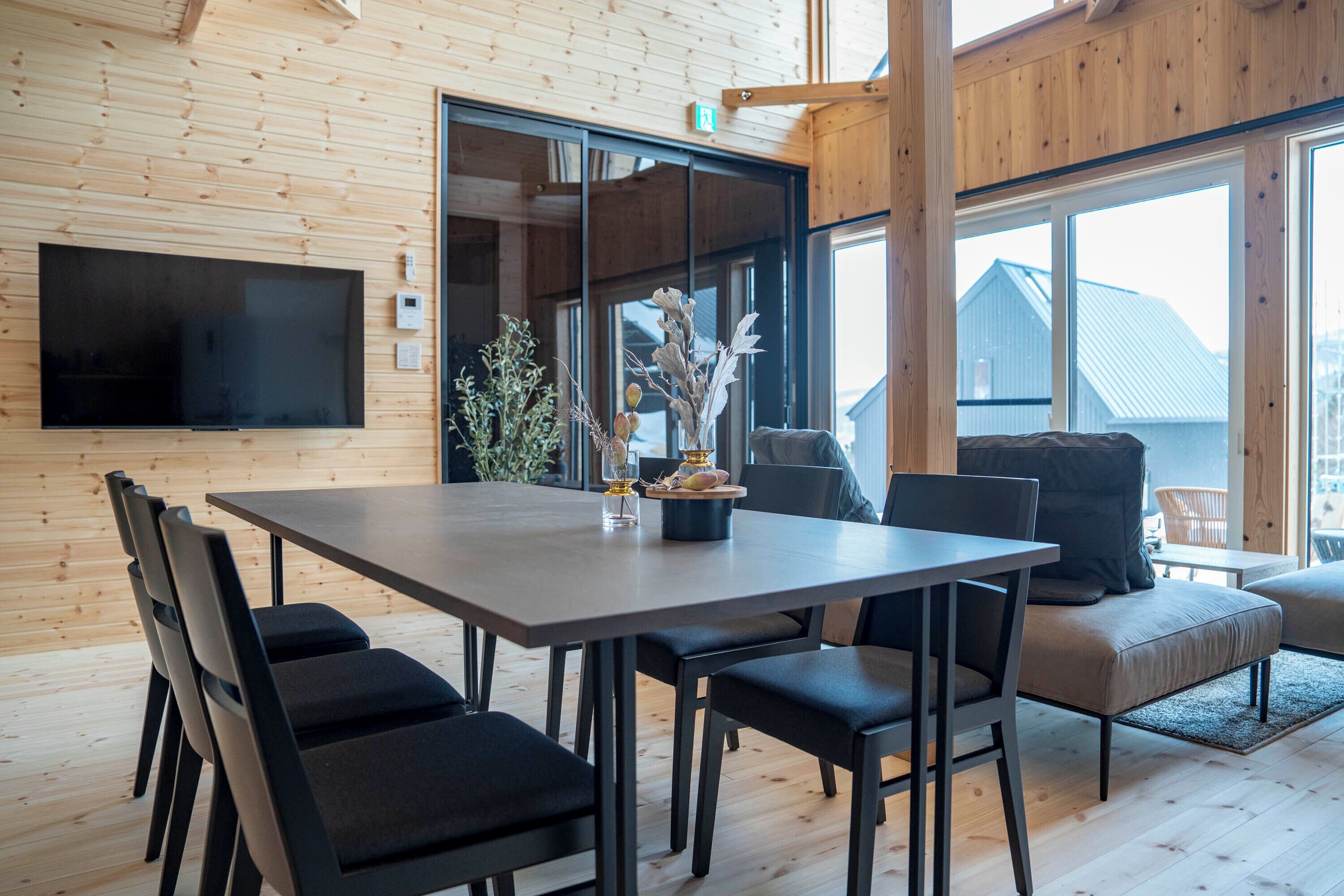 A modern dining area with a large table, black chairs, and a television mounted on the wall.