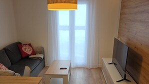 Una sala moderna con un sofá gris, una mesa de centro de madera, un televisor sobre un mueble bajo y un ventanal con cortinas translúcidas.